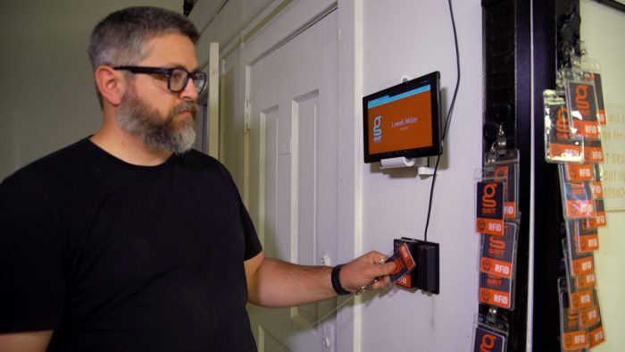 Person demonstrating RFID card scan at wall-mounted user sign in station with touchscreen displaying login confirmation